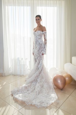 Elegant lace wedding dress with off-shoulder design and flowing train photographed against white curtain backdrop.