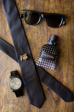 Men's formal accessories including black tie, watch, sunglasses and bracelet arranged on wooden surface.