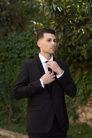 Person in black suit adjusting white tie and collar in outdoor garden setting.