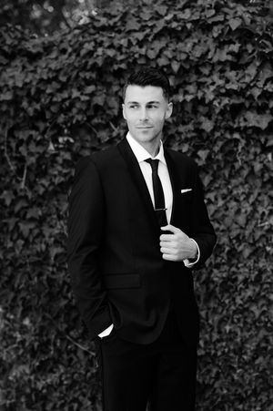 A formal portrait against an ivy wall showing a person in a classic black suit and tie.