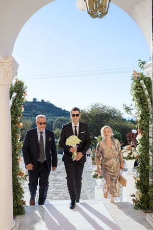 Wedding party walks through Mediterranean archway decorated with greenery and flowers.
