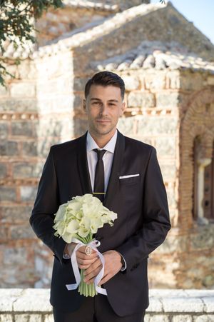 Groom in black suit holding white floral bouquet against stone building backdrop.