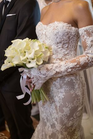 Close-up details of a white lace wedding dress with off-shoulder design and calla lily bouquet at formal ceremony.