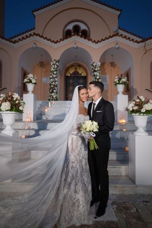 Romantic evening portrait at Mediterranean church entrance with white floral arrangements and candles.