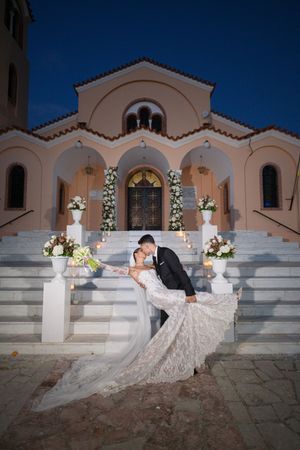 Wedding couple posing on white steps in front of illuminated Mediterranean church at night.