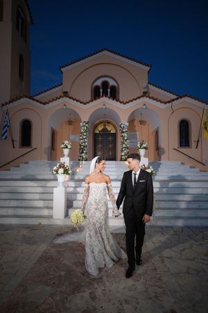 Couple exits white Mediterranean-style church under deep blue evening sky.