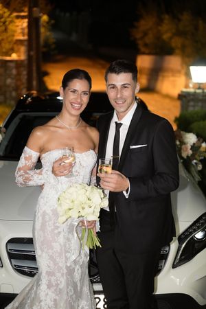 A wedding couple poses next to a white luxury car at night holding a bouquet of white flowers.