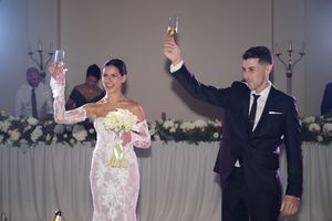 A bride in a white lace gown and groom in a black suit raise champagne glasses during a wedding reception toast.