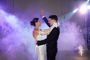 Newlyweds dance together surrounded by purple smoke effects and spotlights during their first dance at a wedding reception.