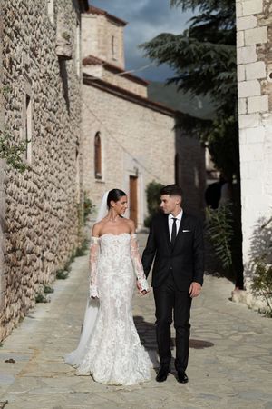 Elegant wedding couple walking along a stone path beside a rustic Mediterranean church building.