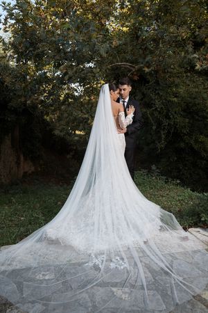 Bride's dramatic lace cathedral veil trails behind on garden path.