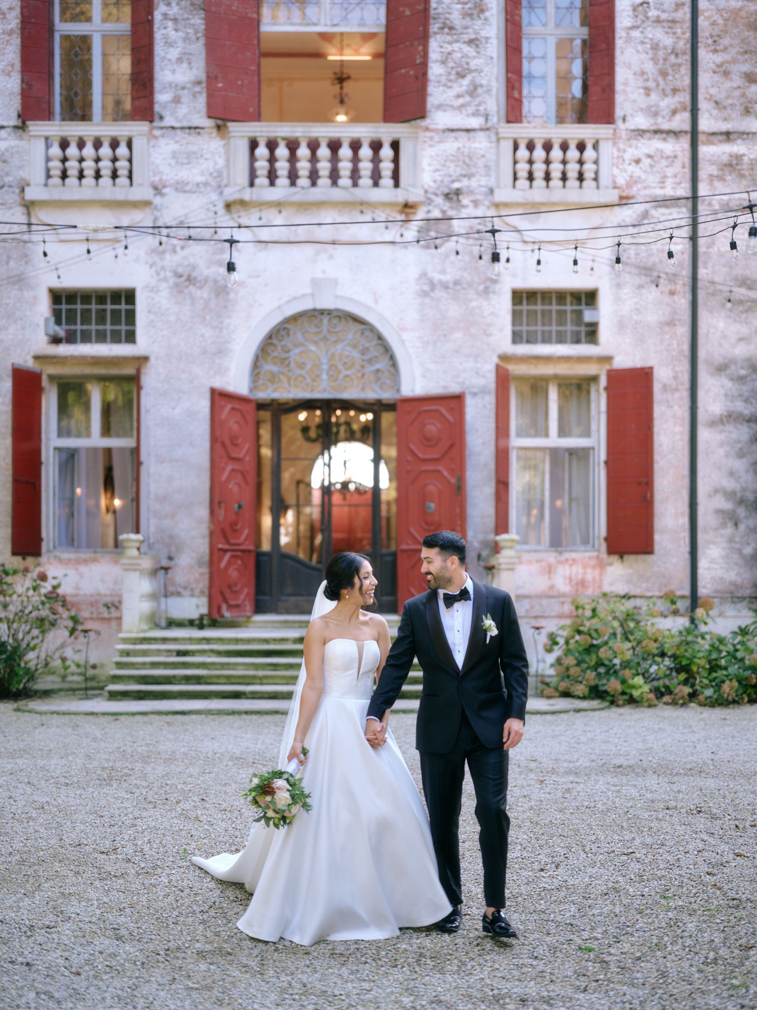 A wedding couple poses for romantic photos in front of a historic villa with red shutters and white walls.