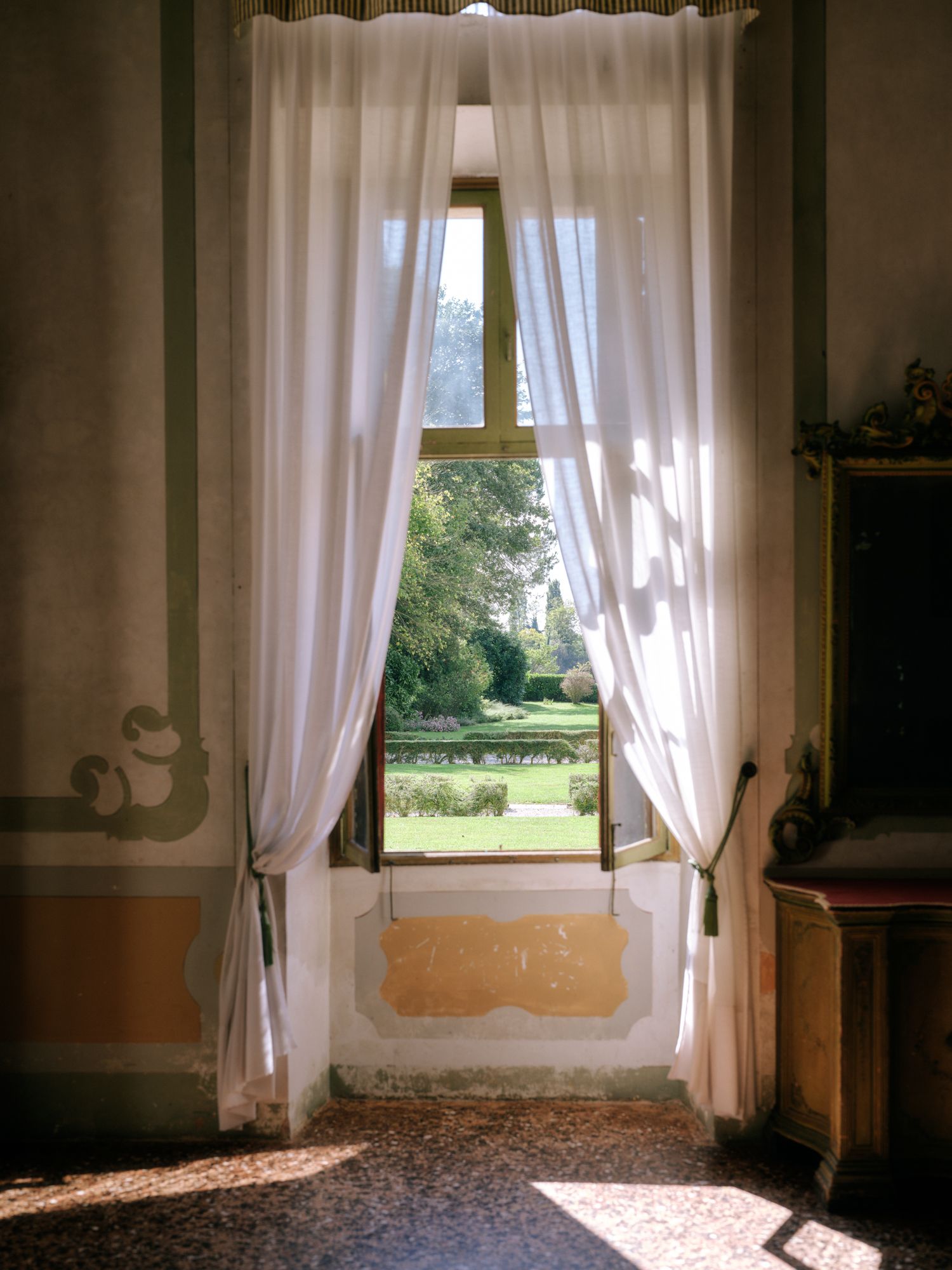 Sunlight streams through sheer white curtains in a row of windows overlooking a garden view.