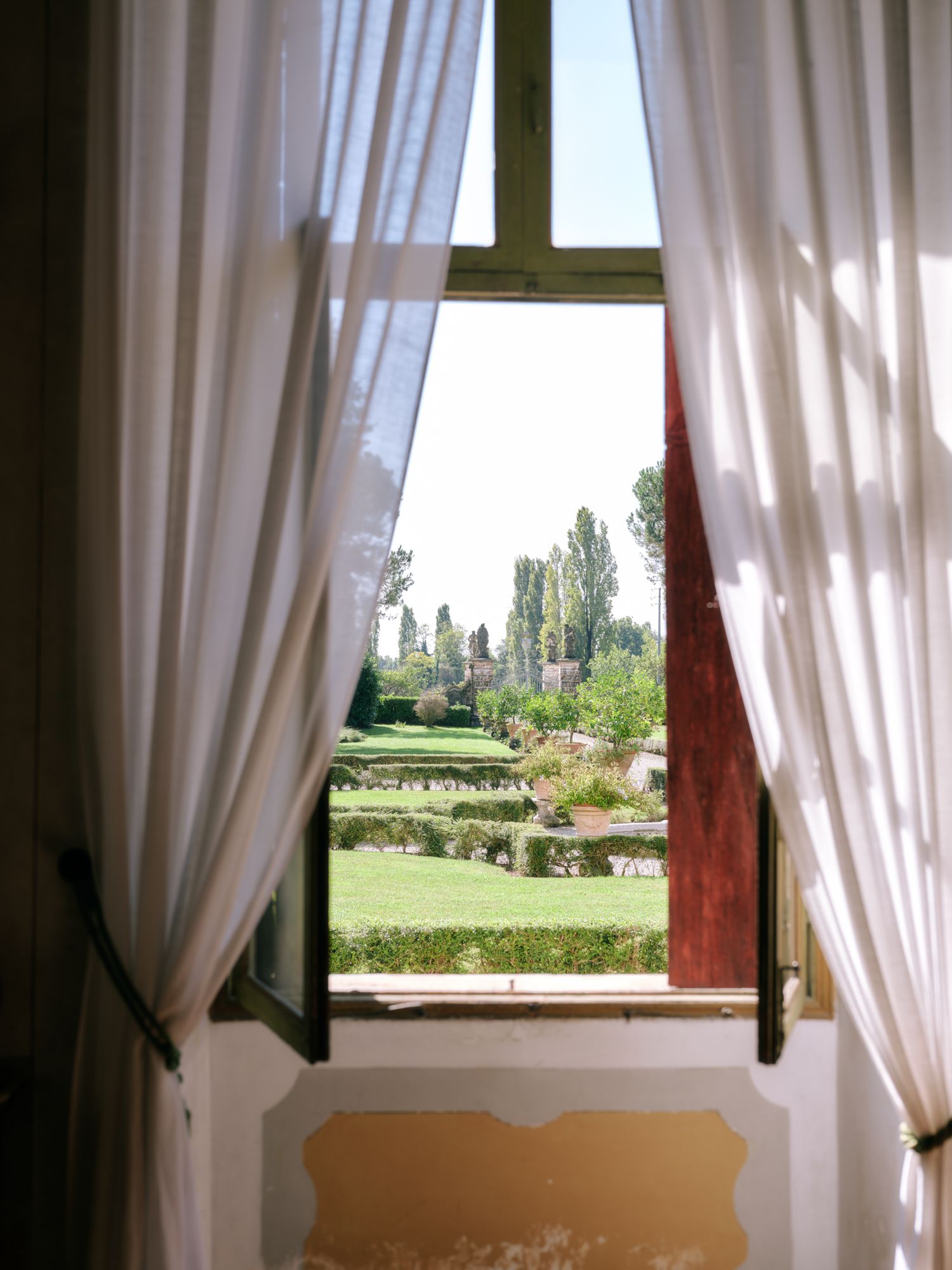 White curtained windows frame a scenic garden view with manicured lawns and hedges.