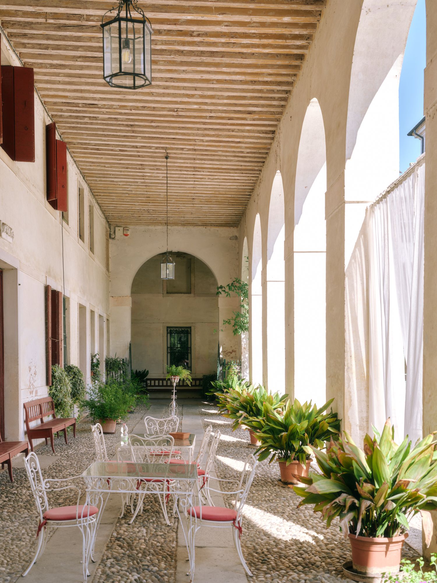 Sunlit Mediterranean-style courtyard with arched colonnade, white wrought iron furniture and potted plants along a covered walkway.