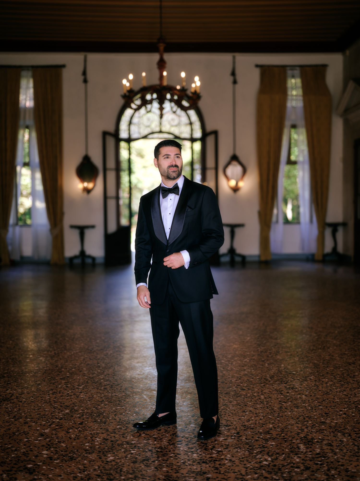 A person in a black suit stands alone in an elegant ballroom with chandelier lighting and ornate architectural details.