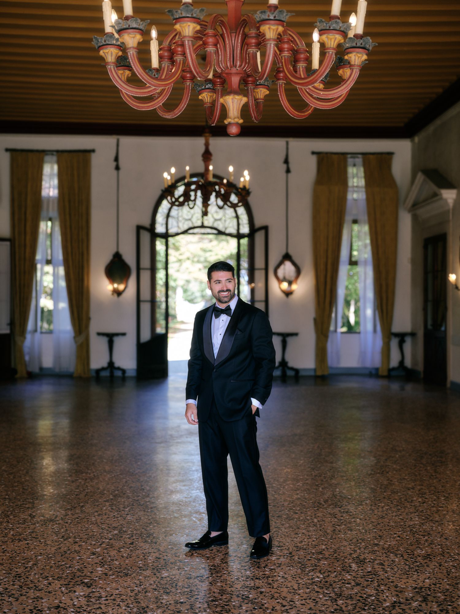 A person in a black tuxedo and bow tie stands in an elegant ballroom with ornate chandeliers and polished floors.