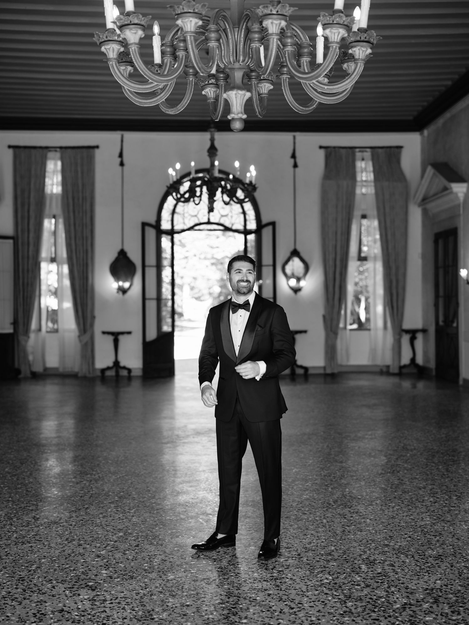 Black and white wedding photos showing groom in tuxedo standing in elegant ballroom with chandelier and hardwood floors.