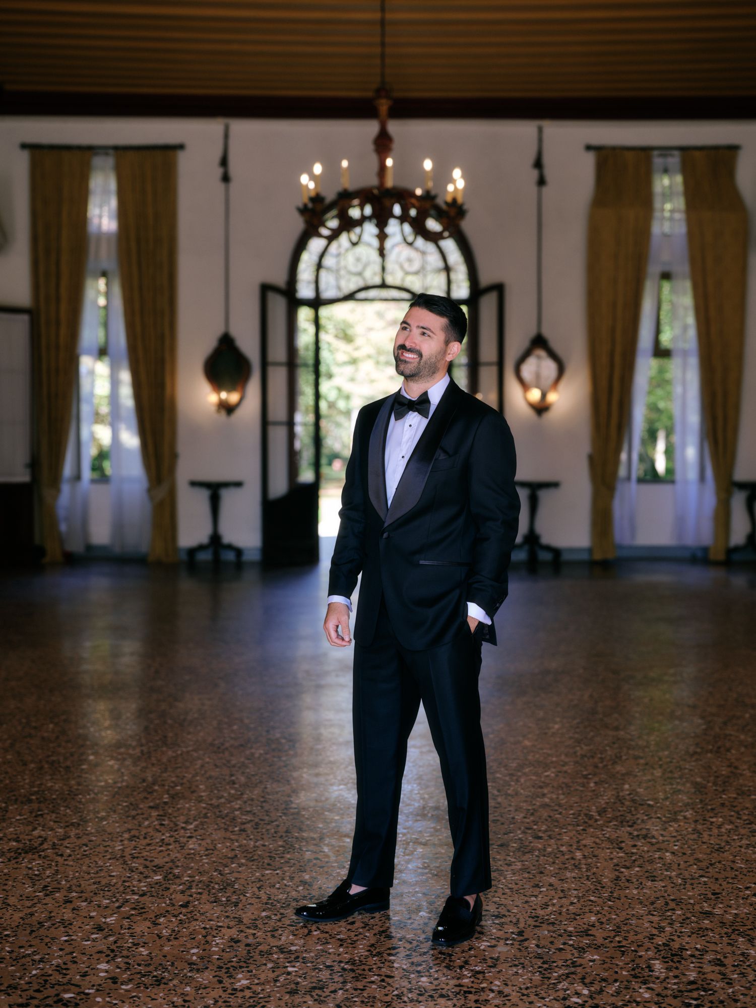 A man in a dark suit poses in a grand ballroom with elegant lighting and wooden floors.