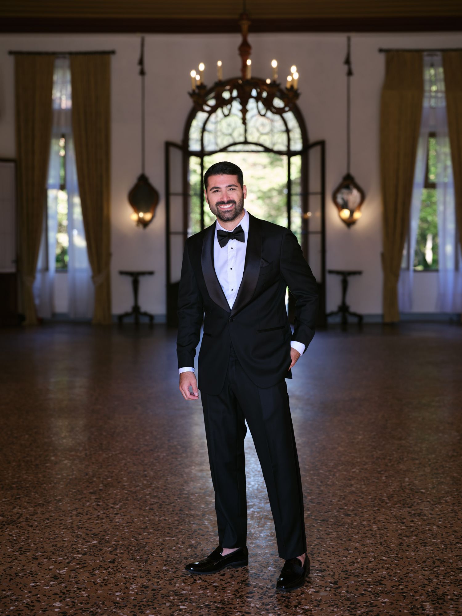 A person in a black tuxedo and bow tie stands in an elegant ballroom with arched windows and a chandelier.
