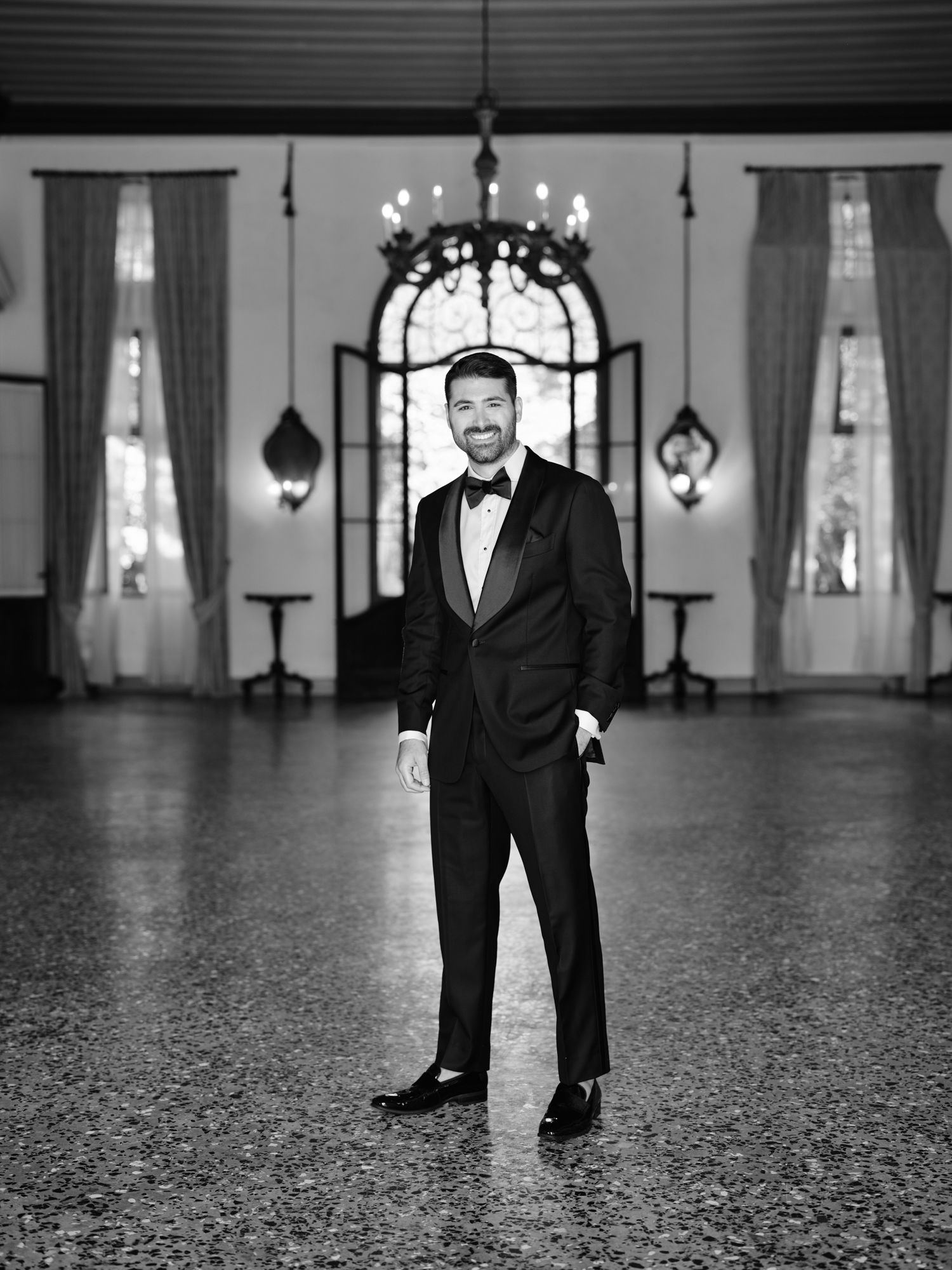 Black and white photo of someone in a tuxedo standing in an elegant ballroom with chandeliers and tall windows.