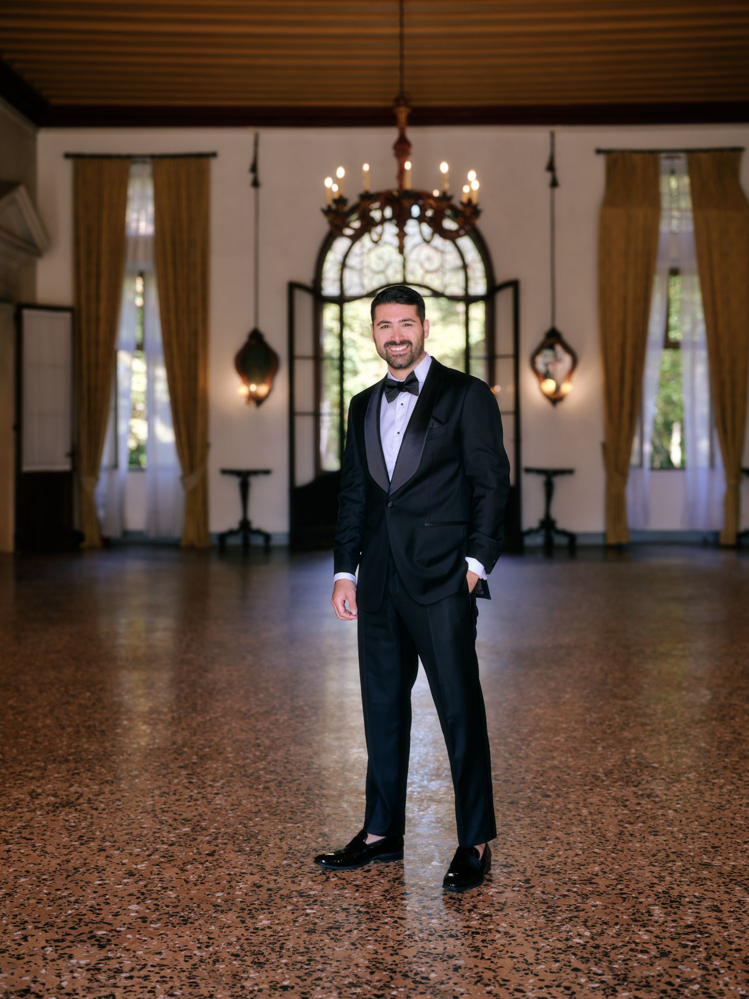 A person in a formal black tuxedo stands on a polished granite floor in an elegant ballroom with chandeliers.