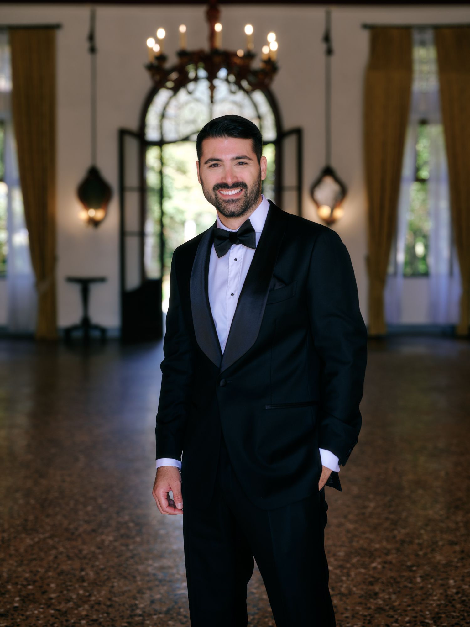 A person in a classic black tuxedo and bow tie smiles while standing in an elegant venue with chandeliers and arched windows.