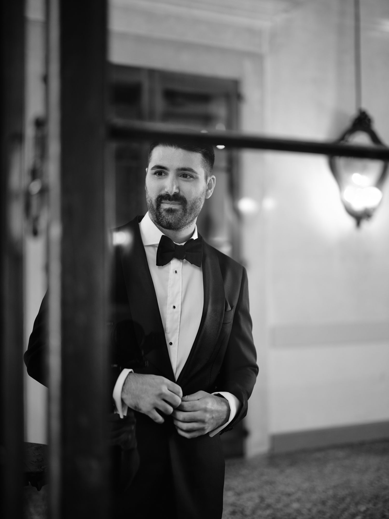 A groom in a black tuxedo and bowtie adjusts his suit jacket in a dimly lit hallway.