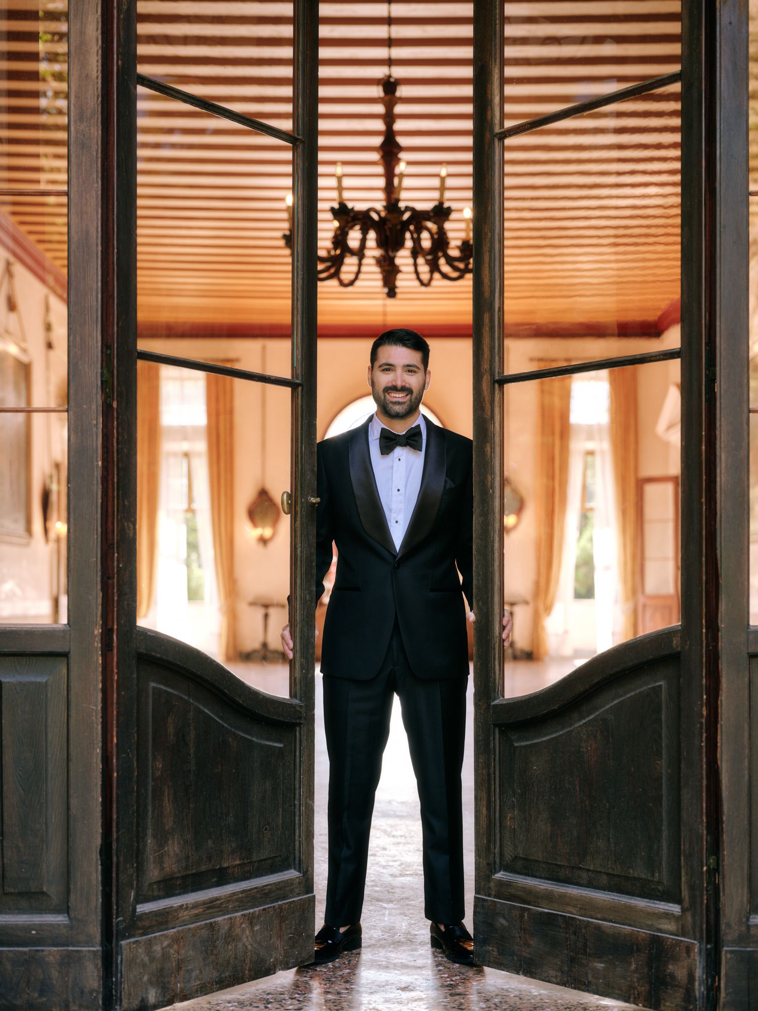 A person in a black tuxedo and bowtie stands in an elegant doorway with warm lighting and wooden details.