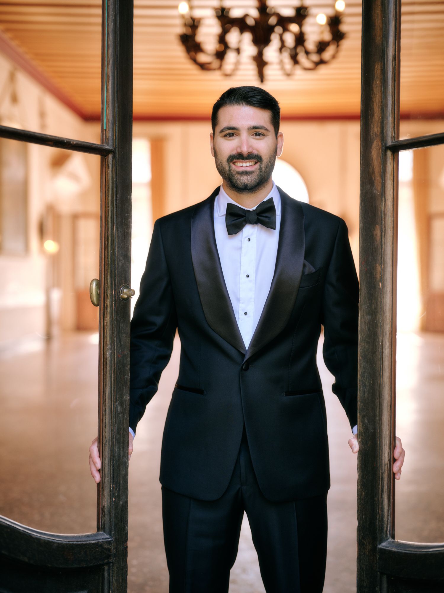 A person in a classic black tuxedo and bowtie poses in an elegant indoor setting with warm lighting.