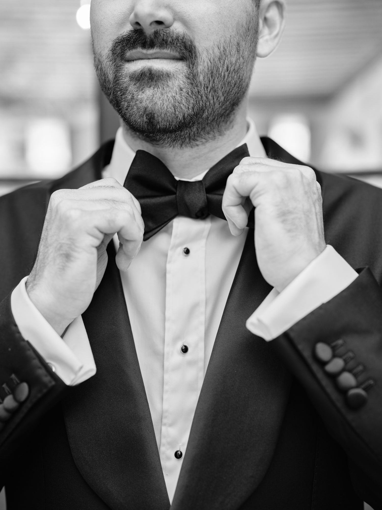 Close-up black and white photo of someone adjusting a bow tie with a tuxedo and white dress shirt.