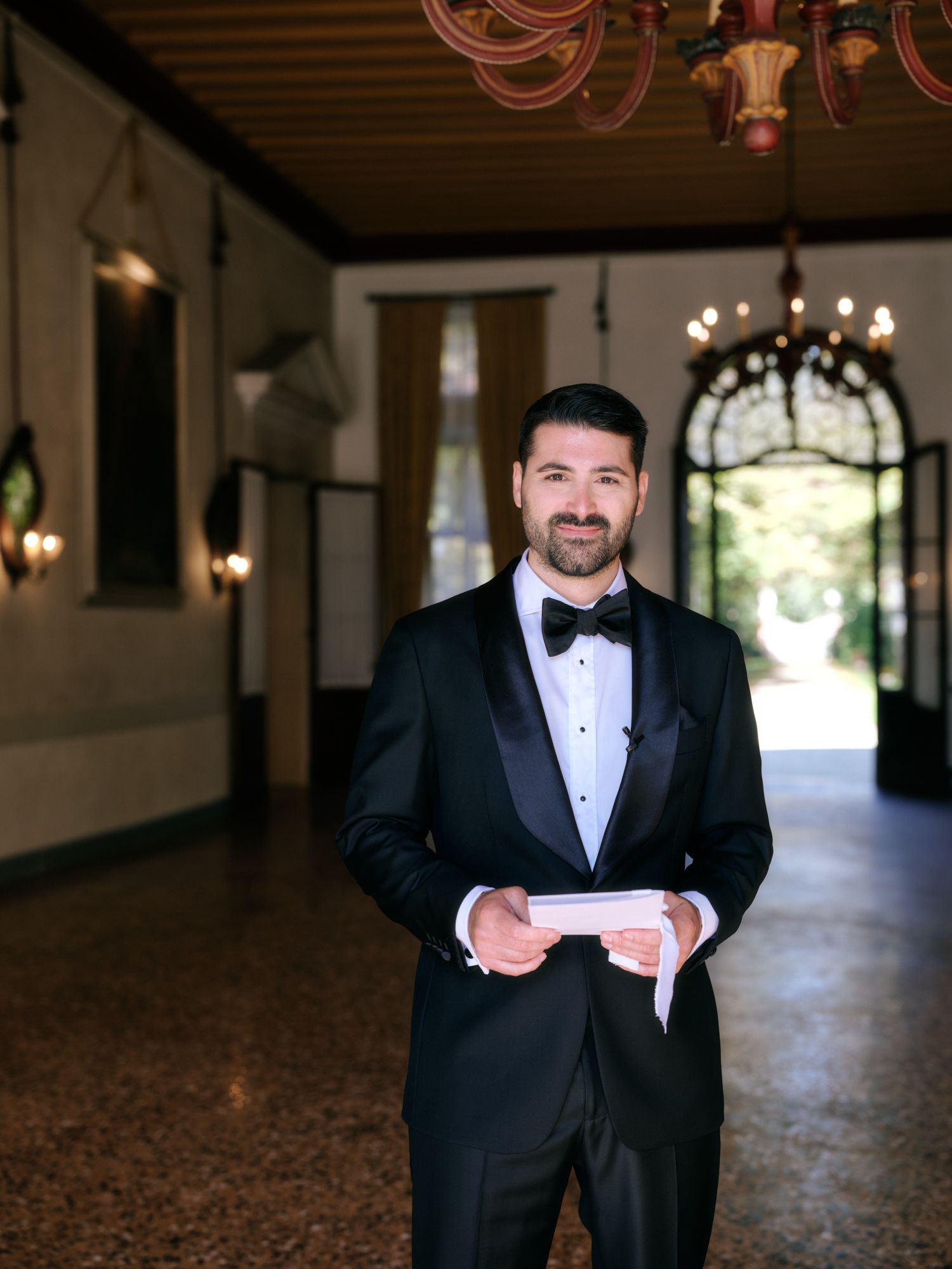 A groom in a black tuxedo and bow tie poses in an elegant venue hallway with chandeliers and patterned carpet.