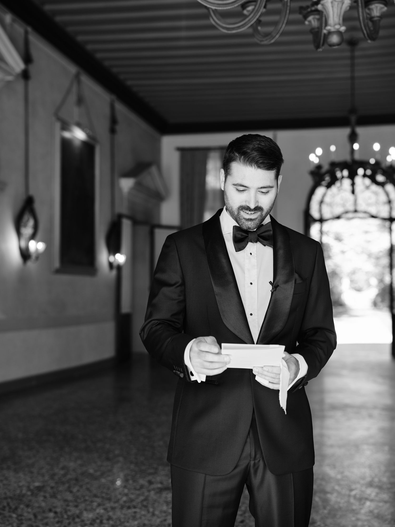 A groom in a classic black tuxedo and bowtie reads from a paper during an emotional wedding ceremony moment.