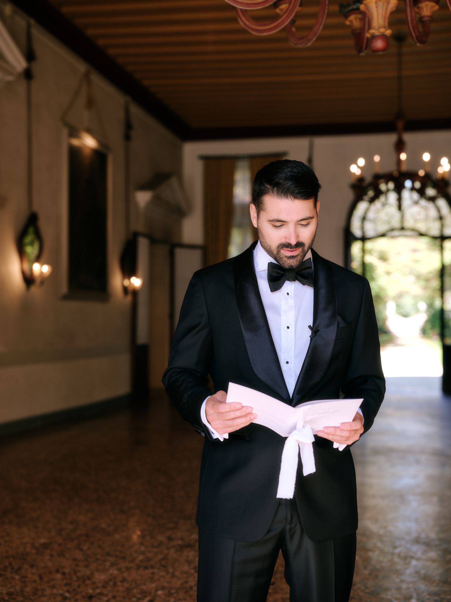 A groom in a black tuxedo and bowtie reads a letter while standing in a dimly lit hallway.