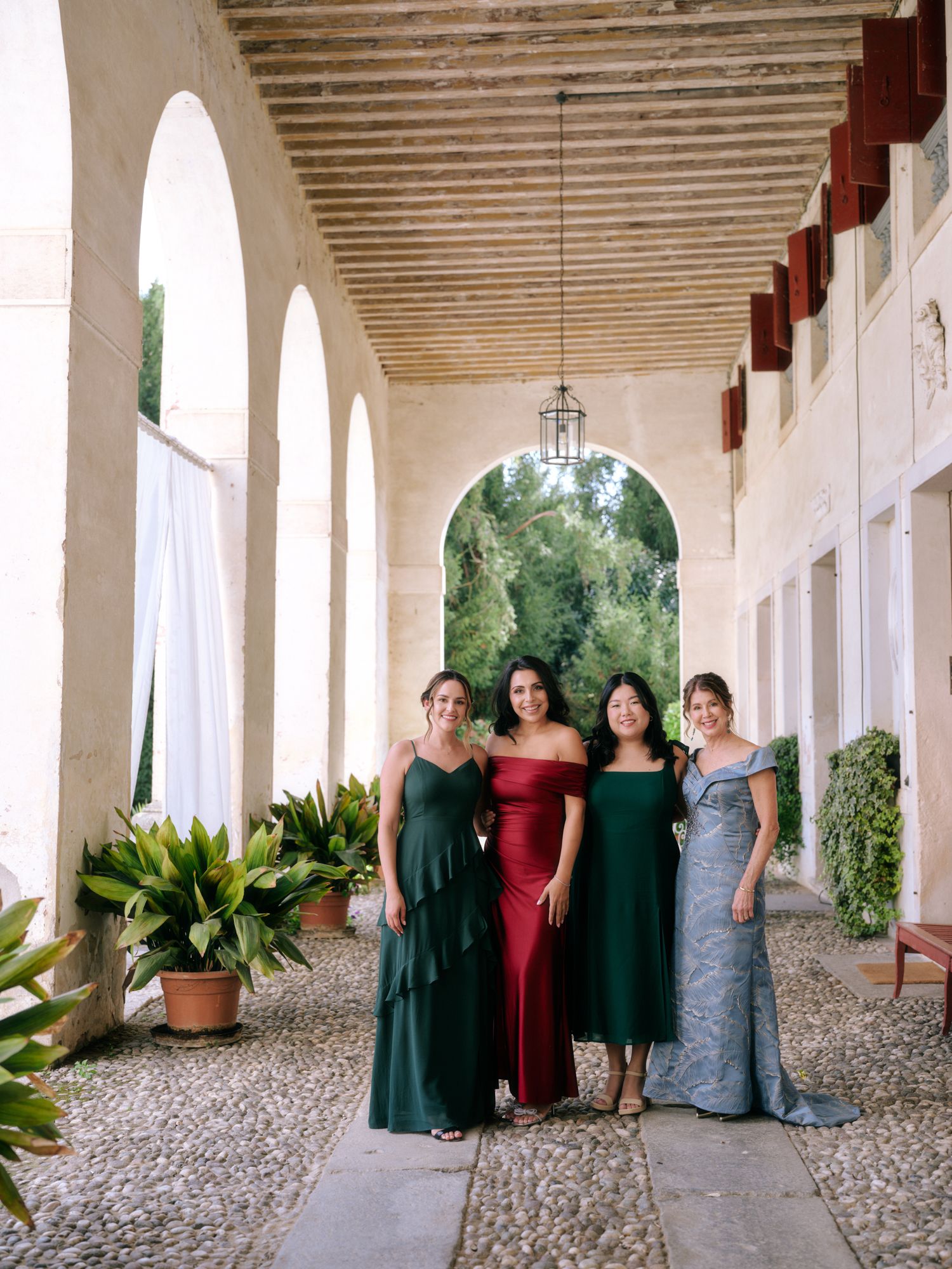 A group in formal attire poses along an arched colonnade walkway with stone columns and gravel floor.