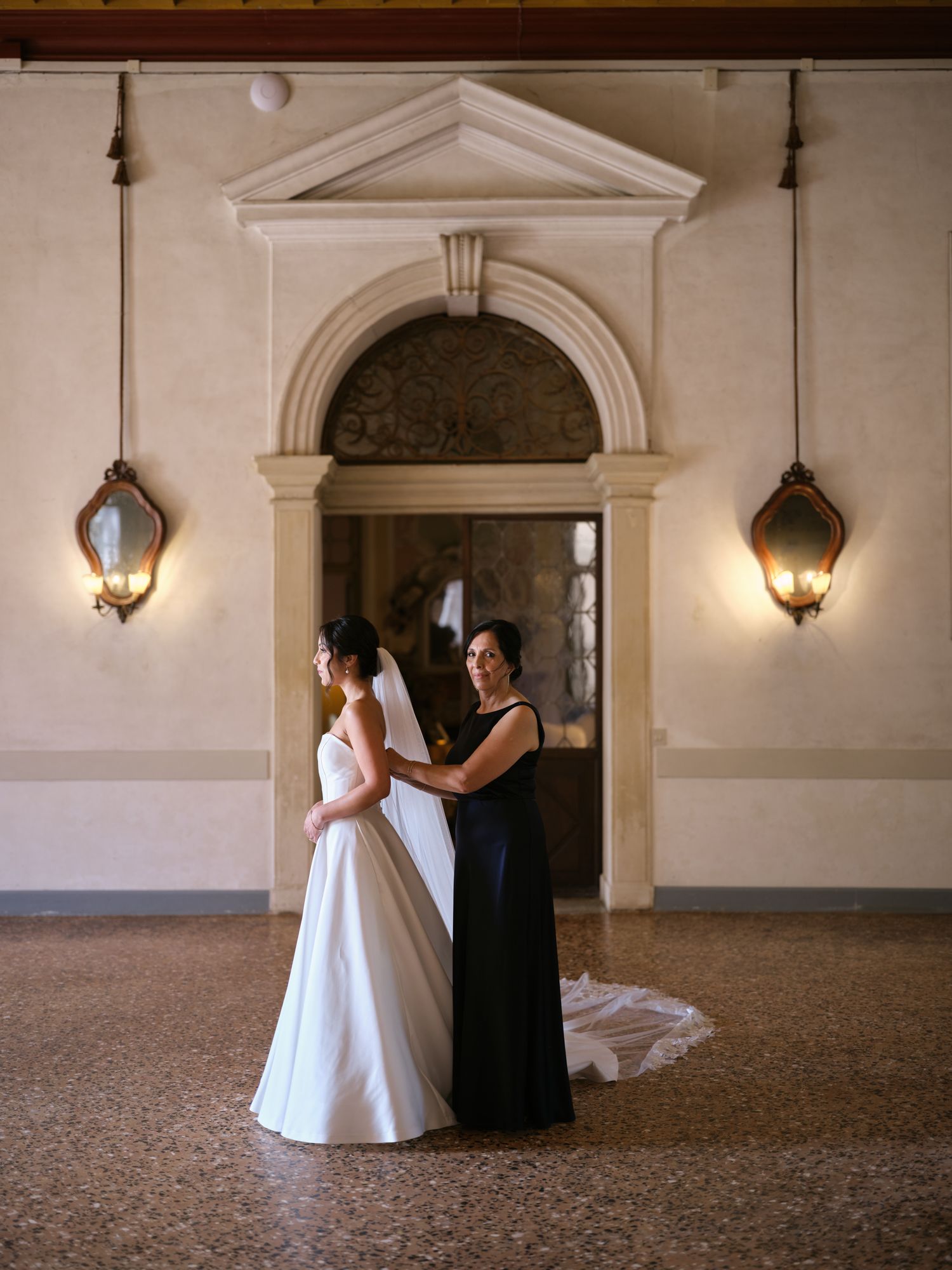 Two figures in contrasting white and navy dresses stand in an elegant hallway with arched doorways and wall sconces.