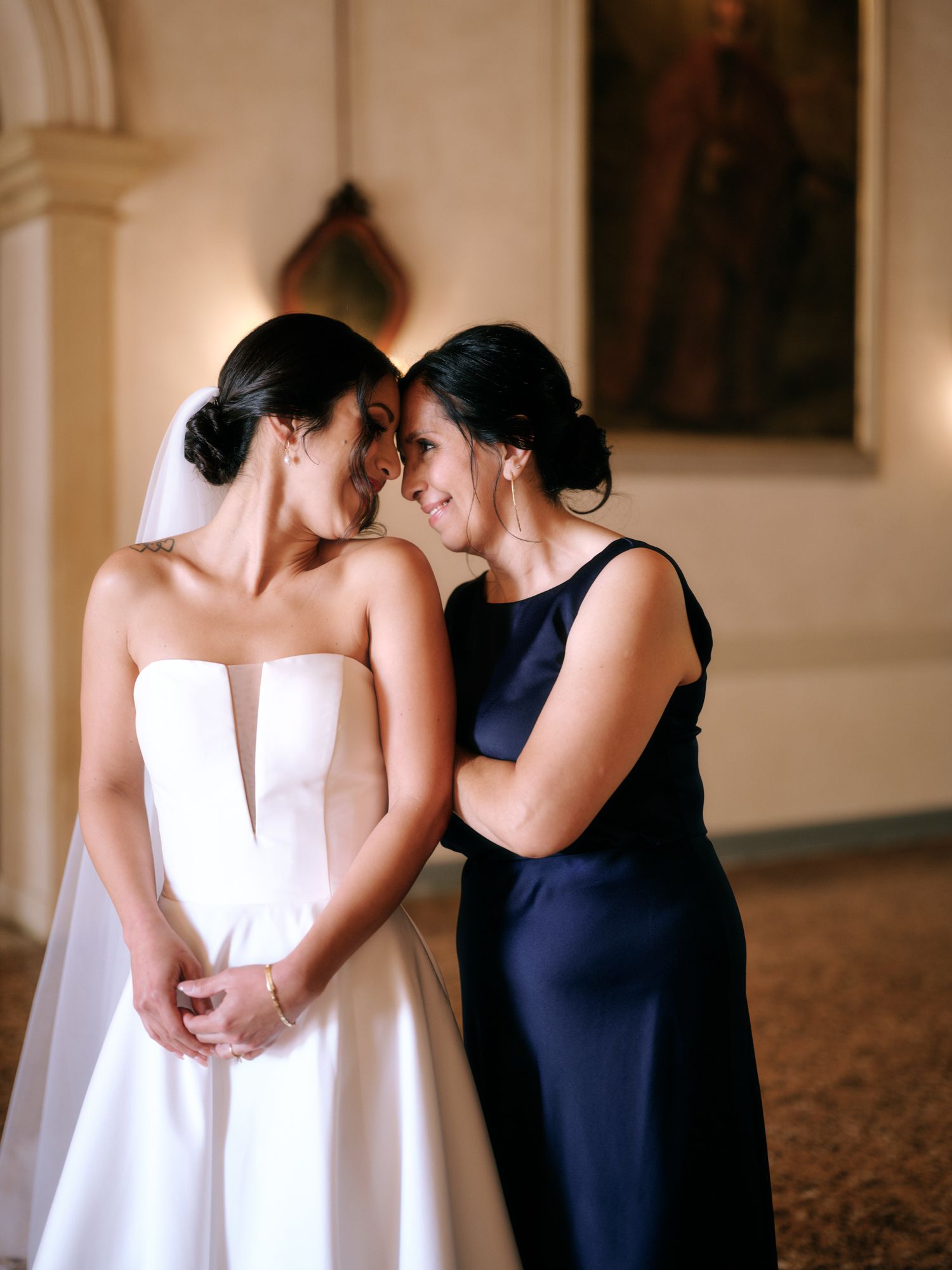 Two women share an intimate moment in formal attire, one wearing a white wedding gown and the other in a navy dress.