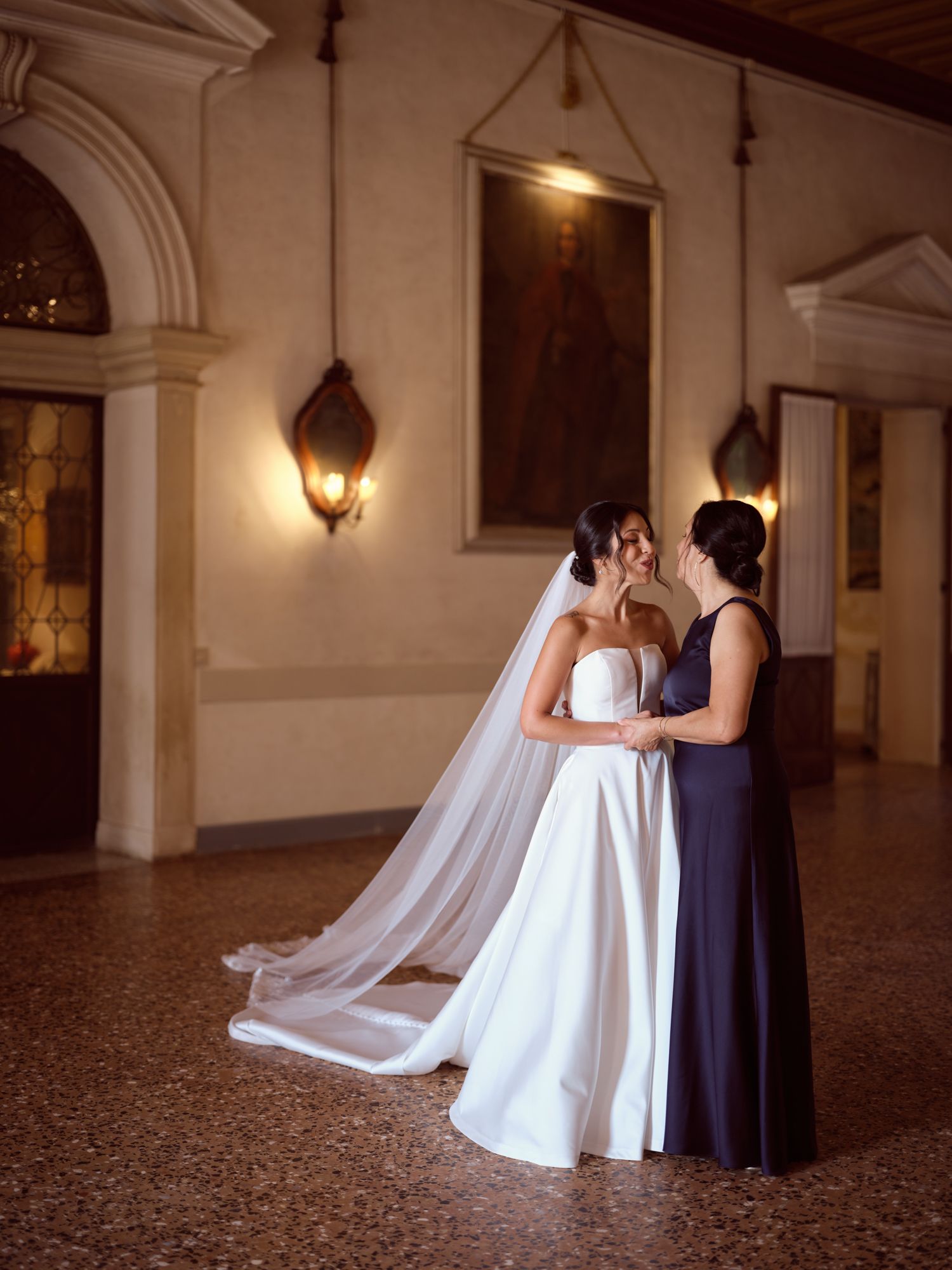 Two people in wedding attire share a romantic dance in an elegant ballroom with warm lighting and ornate columns.