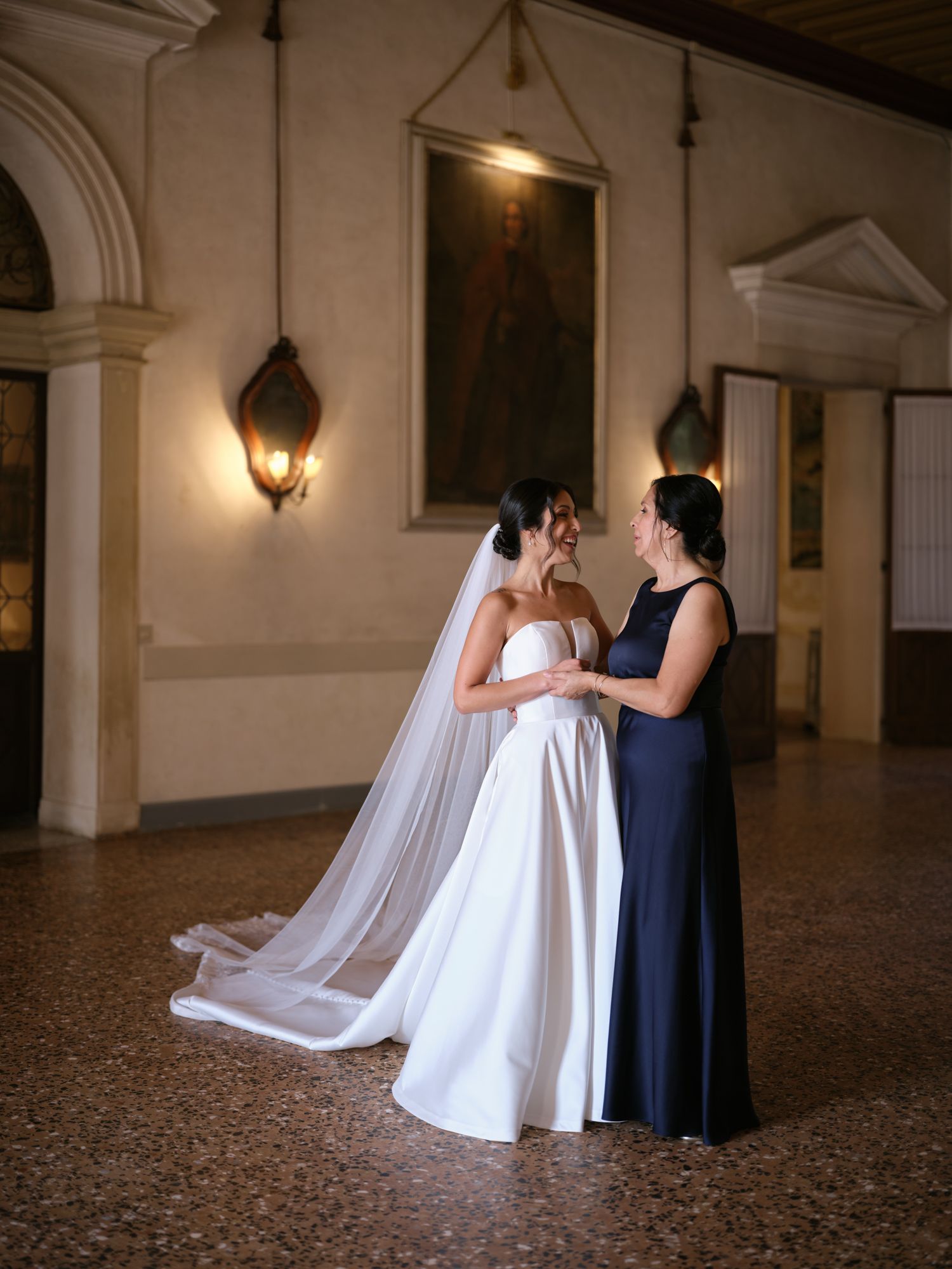 A bride in a white gown and flowing veil stands with someone in a navy dress in an elegant arched hallway.