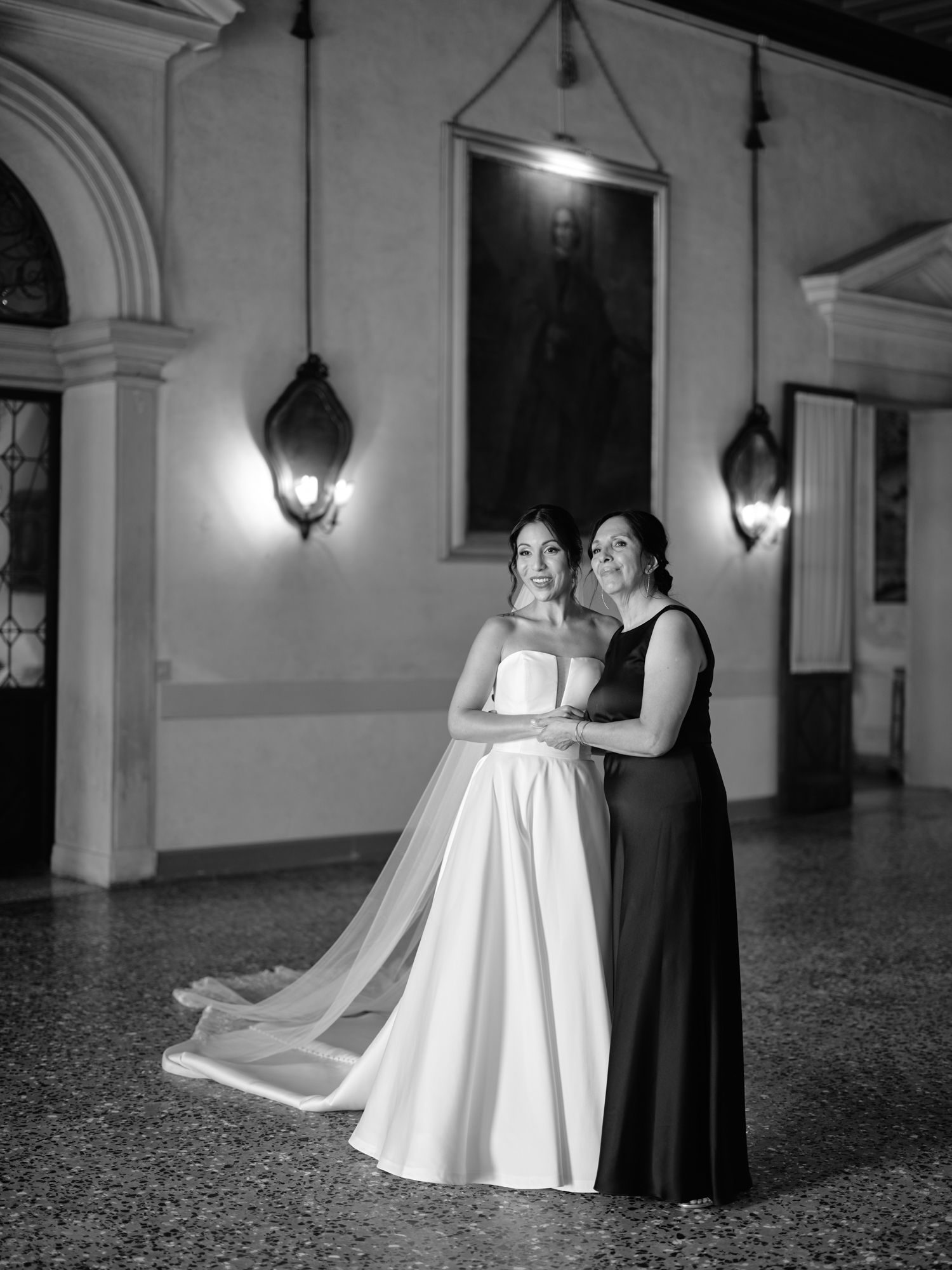 Two elegantly dressed figures pose together in a grand hallway with ornate wall sconces and architectural details.