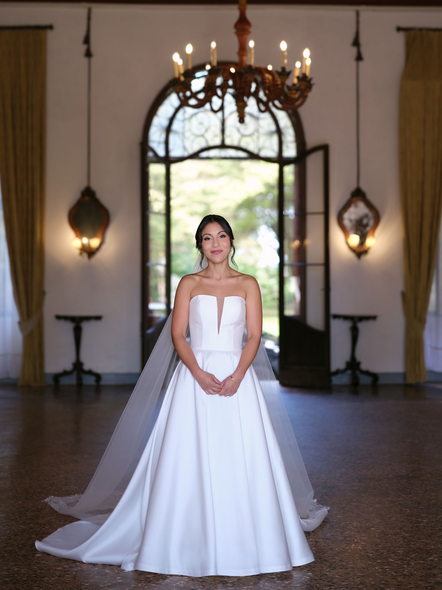 A bride in a white ball gown with cape sleeves poses in an elegant venue with arched windows and chandeliers.