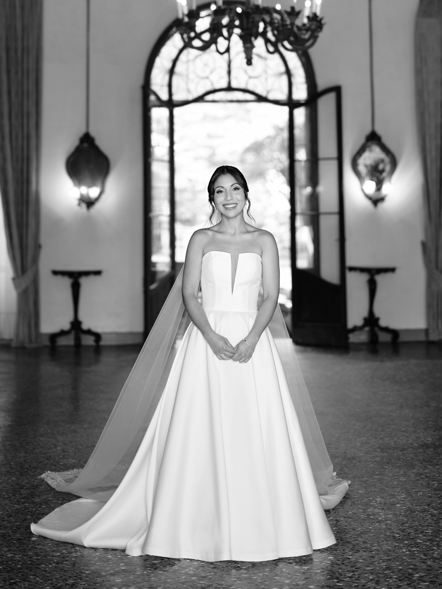 Elegant bride in flowing white ball gown poses in grand hallway with ornate chandeliers and arched windows.