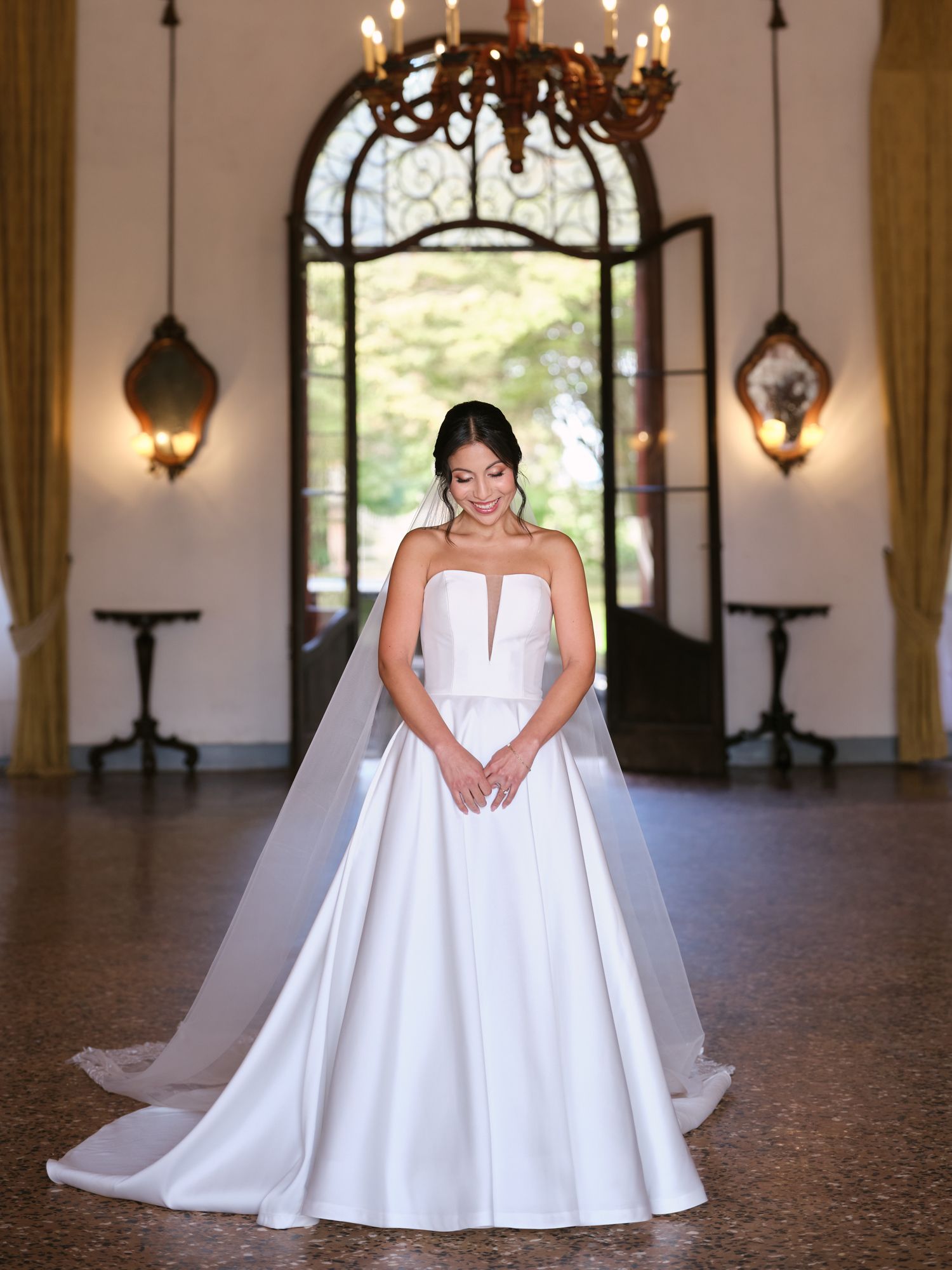 A bride in a stunning white ballgown poses gracefully in an elegant hall with arched windows and chandeliers.