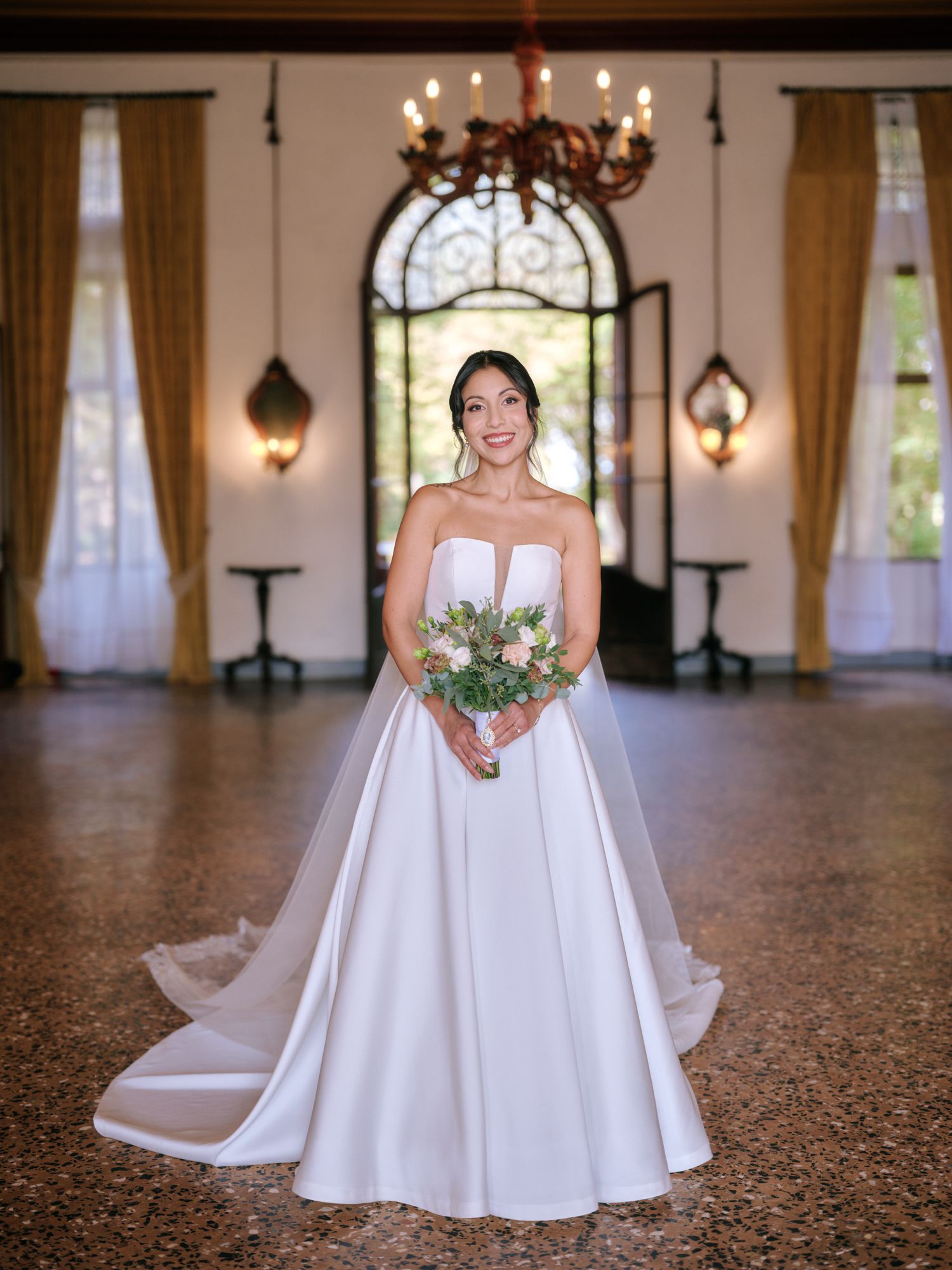 A bride in a classic white strapless ball gown poses in an elegant venue with arched windows and chandeliers.
