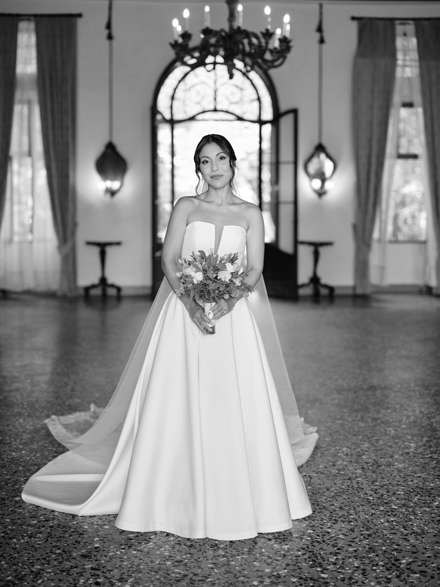 Black and white photo of a bride in a strapless ball gown holding a bouquet in a grand hall with chandeliers.