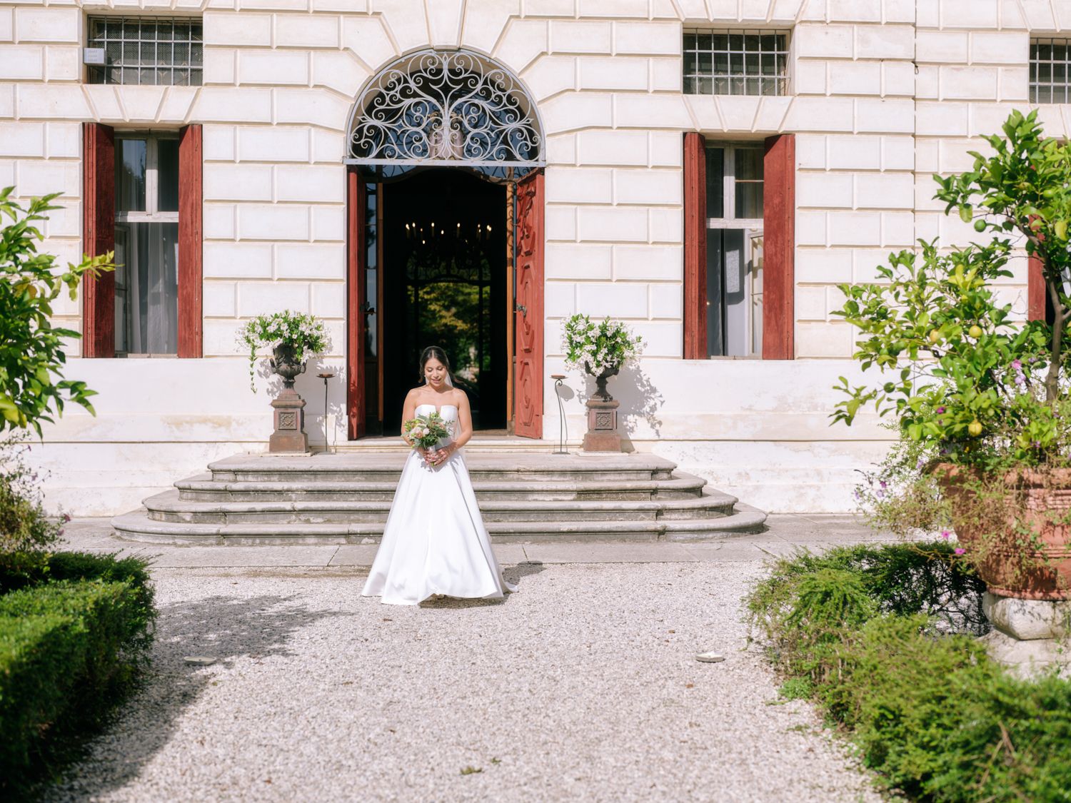 A historic building exterior with stone steps, arched windows, and pink-tinted walls in a formal garden setting.