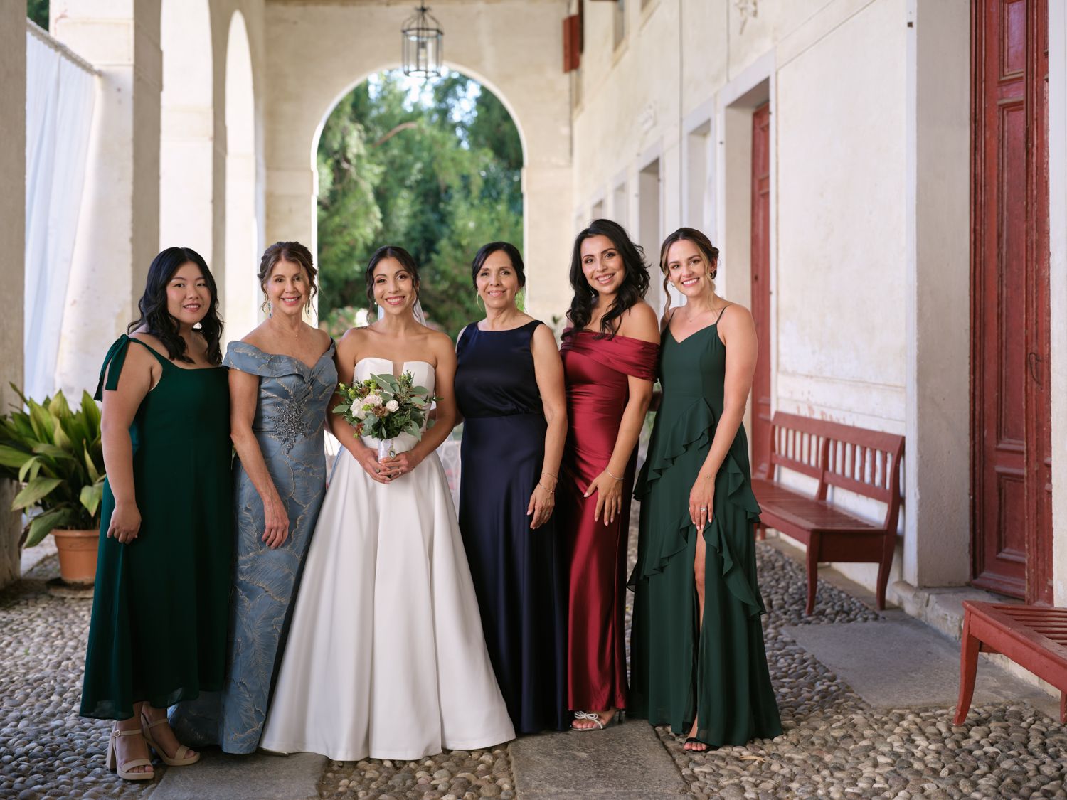 Wedding party members in dark green and black dresses pose together in an outdoor courtyard setting.