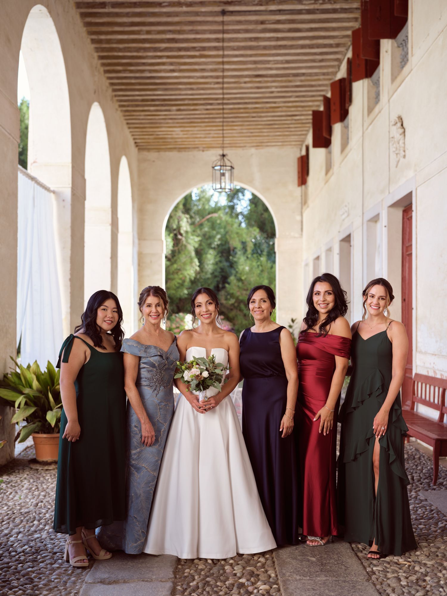 A wedding party poses together in an elegant covered walkway with white columns and rustic wooden ceiling beams.