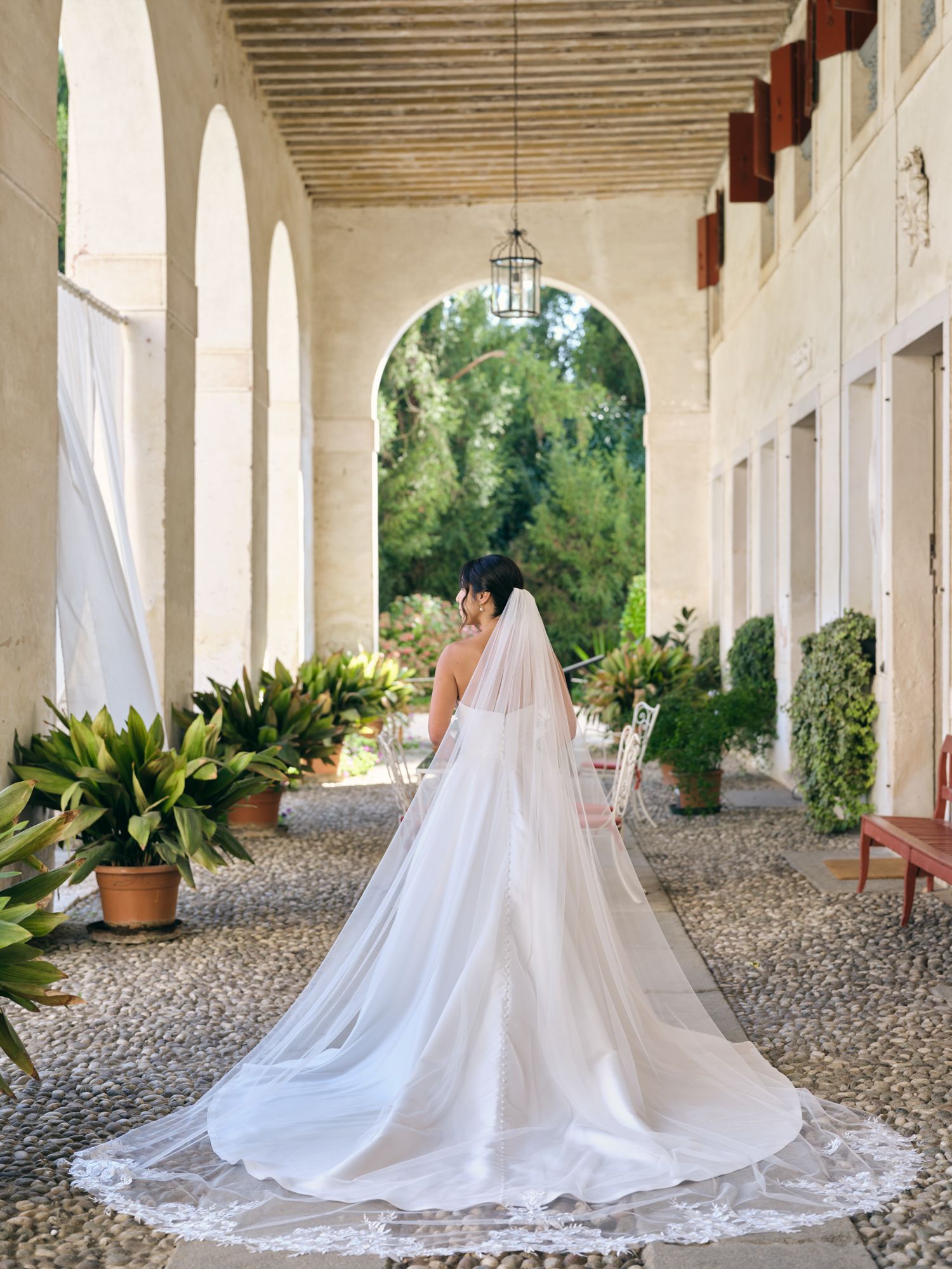 A flowing white wedding gown and veil photographed from behind in an elegant arched colonnade with potted plants.