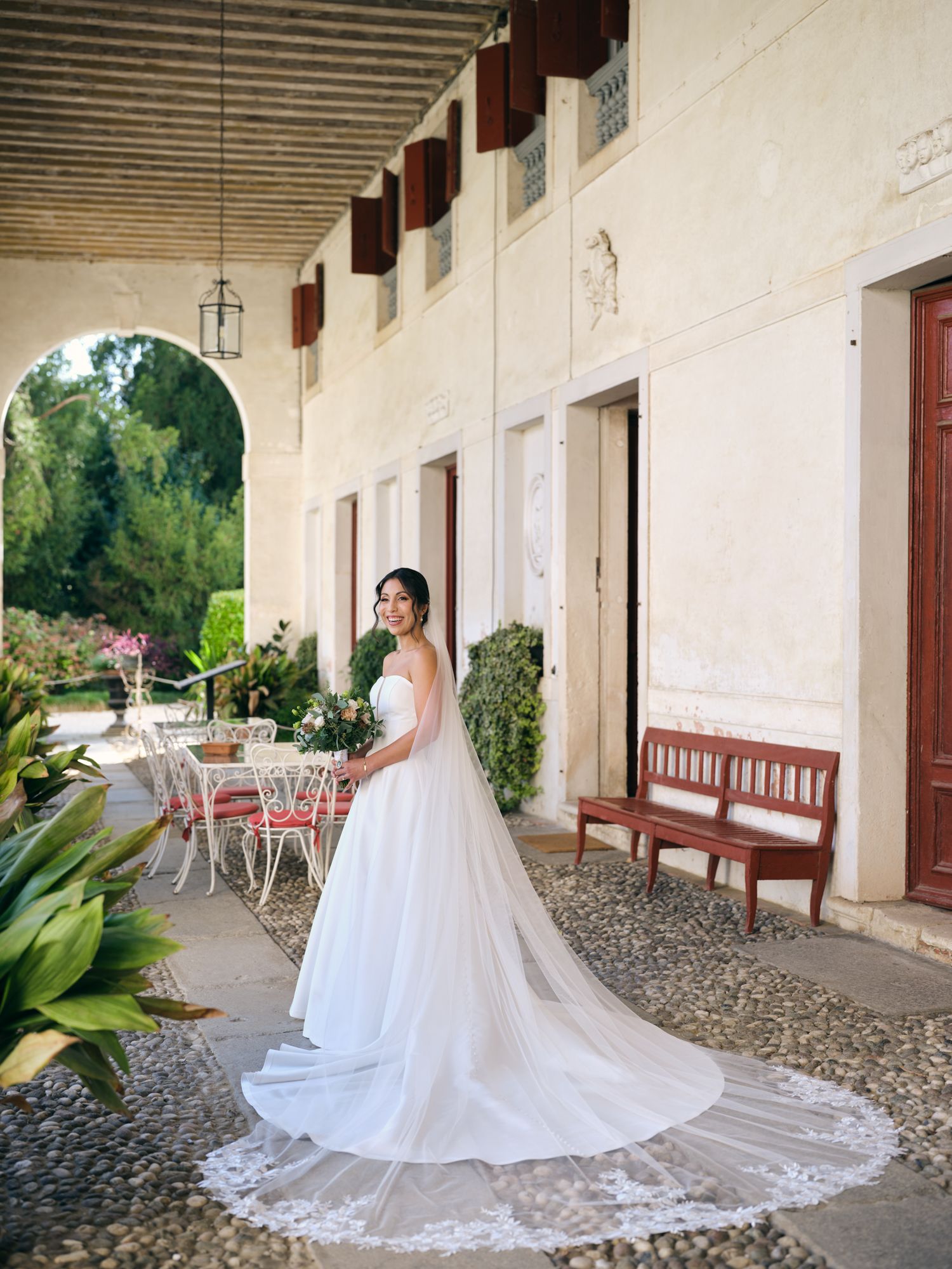 A bride in a flowing white wedding gown and cathedral veil poses along a rustic covered walkway with stone columns.